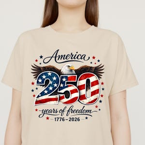 May include: Beige t-shirt with a patriotic design. The graphic includes the word "America", the number "250" in red, white, and blue, an eagle, and the phrase "years of freedom" with the years 1776-2026.