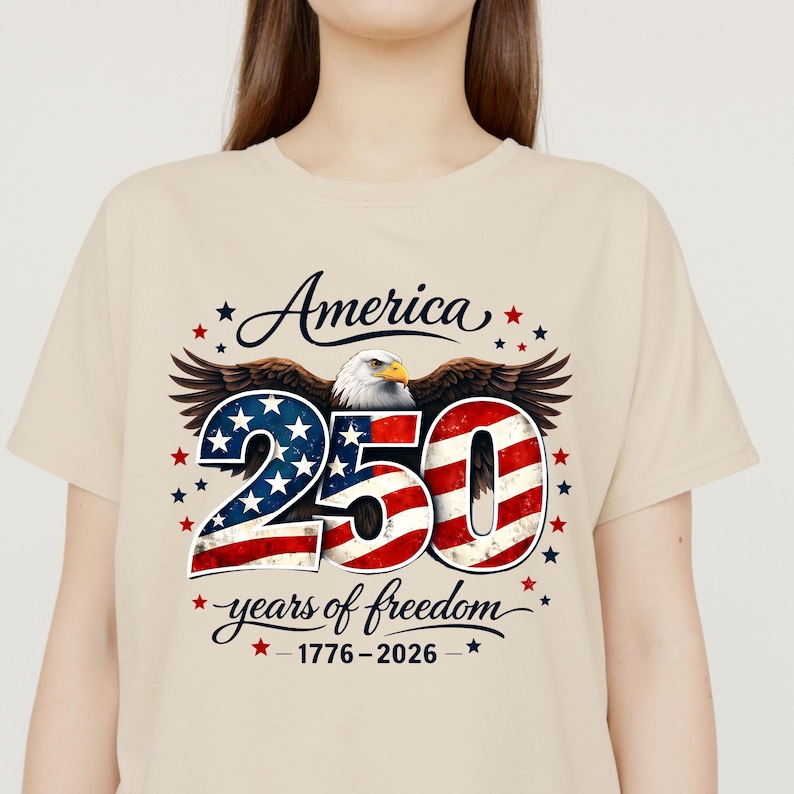 May include: Beige t-shirt with a patriotic design. The graphic includes the word "America", the number "250" in red, white, and blue, an eagle, and the phrase "years of freedom" with the years 1776-2026.