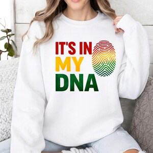 May include: White sweatshirt featuring the text "IT'S IN MY DNA" in red, yellow, and green. A fingerprint design in the same colours is to the right of the text. The sweatshirt has a crew neck.
