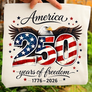 May include: A cream tote bag with the word "America" and an eagle illustration. The number "250" is designed with the American flag, and the text "years of freedom" and dates "1776-2026" are below. The bag is held by a person.
