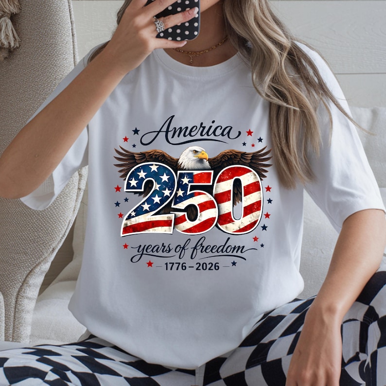 May include: White t-shirt with a patriotic design celebrating 250 years of American freedom. The design features an eagle, the word "America", and the number "250" in red, white, and blue, with the years 1776-2026.