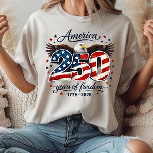 May include: A cream-coloured t-shirt with a patriotic design. The graphic features the word "America" above a stylized eagle. Below the eagle is the number "250" in red, white, and blue, with the text "years of freedom" and the dates 1776-2026.