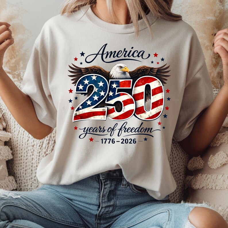 May include: A cream-coloured t-shirt with a patriotic design. The graphic features the word "America" above a stylized eagle. Below the eagle is the number "250" in red, white, and blue, with the text "years of freedom" and the dates 1776-2026.