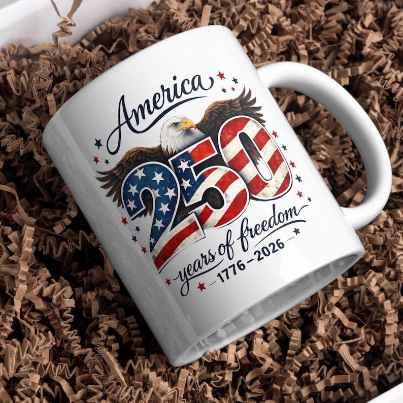 May include: White ceramic mug with a patriotic design. The mug features the word "America" in script, an eagle, and the number "250" in red, white, and blue, with the text "years of freedom 1776-2026".