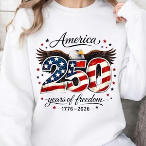 May include: White sweatshirt with a graphic celebrating America's 250th anniversary. The design features an eagle, the word "America", the number "250" in red, white, and blue, and the text "years of freedom 1776-2026".