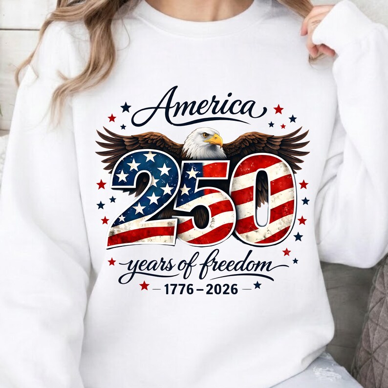 May include: White sweatshirt with a graphic celebrating America's 250th anniversary. The design features an eagle, the word "America", the number "250" in red, white, and blue, and the text "years of freedom 1776-2026".
