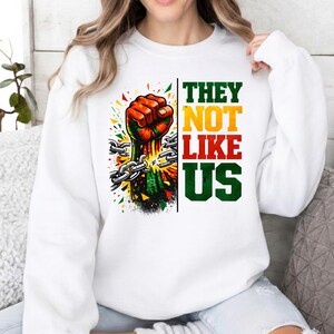 May include: White sweatshirt with a graphic of a clenched fist breaking chains, with colourful accents. The text "THEY NOT LIKE US" is vertically aligned in green, yellow, and red. A powerful statement.