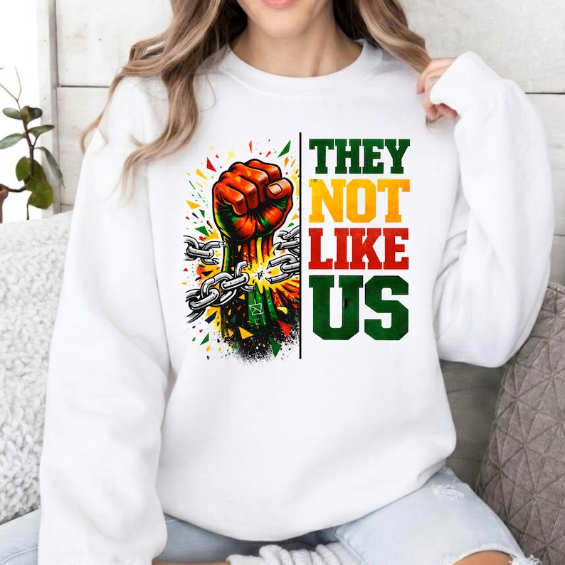 May include: White sweatshirt with a graphic of a clenched fist breaking chains, with colourful accents. The text "THEY NOT LIKE US" is vertically aligned in green, yellow, and red. A powerful statement.