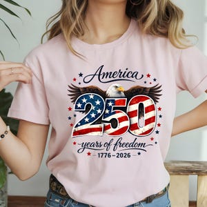 May include: A light pink t-shirt with a patriotic design. The graphic includes the word "America" above the number "250" with stars and stripes, an eagle, and the text "years of freedom - 1776-2026".