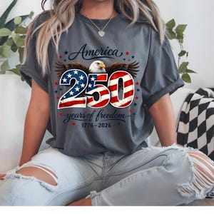 May include: Dark grey t-shirt with a patriotic design celebrating 250 years of American freedom. The design features an eagle, the number 250 in red and white stripes, and the text "America" and "years of freedom". Stars and the years 1776-2026 are also included.