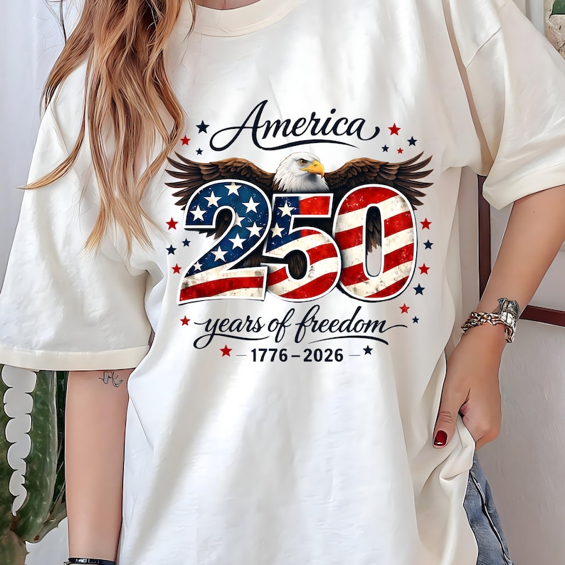 May include: Cream-coloured t-shirt with a patriotic design. The graphic features an eagle, the word "America", the number "250" in red and blue, and the text "years of freedom 1776-2026". Red and blue stars surround the design.