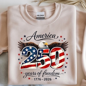 May include: A beige Gildan heavy cotton sweatshirt with the text "America 250 years of freedom 1776-2026". The design features a bald eagle, stars, and the number 250 in a red, white, and blue American flag design.