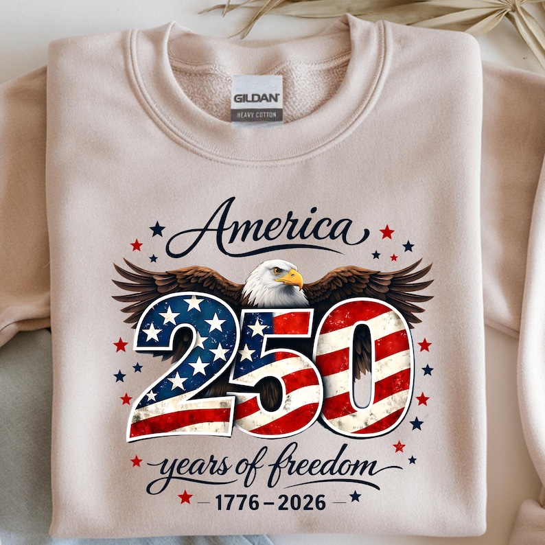 May include: A beige Gildan heavy cotton sweatshirt with the text "America 250 years of freedom 1776-2026". The design features a bald eagle, stars, and the number 250 in a red, white, and blue American flag design.