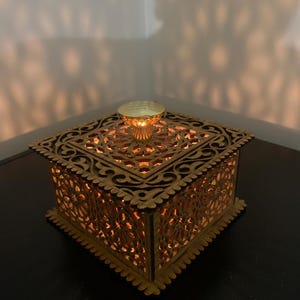 Può includere: Una scatola di legno con intricati intagli e una manopola dorata sul coperchio. La scatola è illuminata dall'interno, proiettando un bagliore caldo.