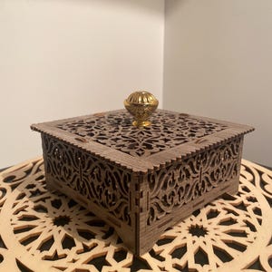 Può includere: Una scatola quadrata in legno con intricati disegni ritagliati e una manopola dorata sul coperchio. La scatola è appoggiata su una superficie in legno con un design ritagliato simile.