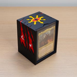 May include: A black rectangular card storage box featuring a yellow eight-pointed star symbol with a red eye in the centre on the top lid. The sides display red claw mark cutouts. A trading card labelled Abaddon the Despoiler is visible.