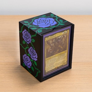 May include: A black rectangular card storage box featuring purple rose illustrations with green vines on the top and side panels. The front of the box displays a single collectible trading card titled The Swarmlord in a clear protective sleeve.