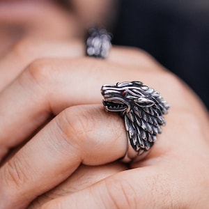 Puede incluir: Un anillo de metal plateado con un diseño detallado de cabeza de lobo, texturas de pelaje y pequeños acentos rojos en los ojos. El anillo presenta una expresión de boca abierta y se asienta de forma prominente con un acabado metálico.