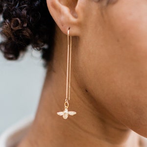 May include: A woman wearing a white shirt with a gold bee charm dangling from a long gold chain earring.