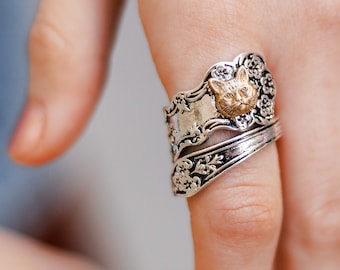 May include: A silver-toned ring with a detailed floral design and a gold-colored cat face. The ring is worn on a finger, showcasing the intricate craftsmanship and unique style of the jewelry.