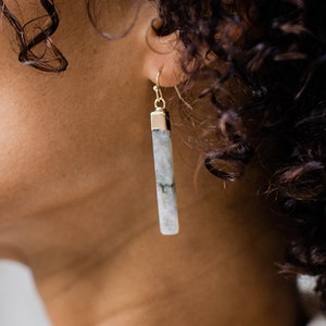 Puede incluir: Primer plano de una persona que lleva pendientes largos y rectangulares. Los pendientes tienen una parte superior dorada y un cuerpo de piedra gris claro y blanco. Los pendientes cuelgan del lóbulo de la oreja.