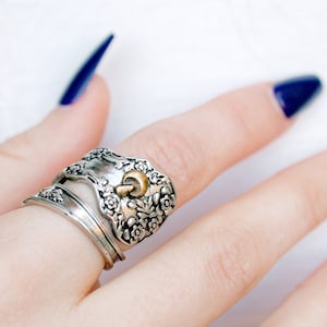 Puede incluir: Un anillo de plata con un diseño floral y un acento dorado. El anillo se lleva en un dedo con esmalte de uñas azul oscuro.