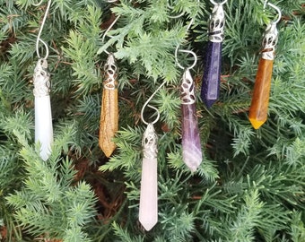 Op de afbeelding: Een verzameling van zeven edelsteenhangers met zilverkleurige metalen doppen en ophangdraden. De edelstenen zijn in verschillende kleuren, waaronder wit, bruin, paars, blauw, roze en geel. De hangers worden gepresenteerd tegen een achtergrond van groen gebladerte.