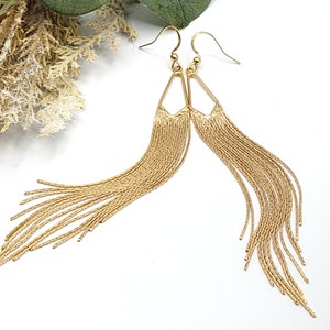 May include: Gold dangle earrings with a geometric triangle design and a long chain fringe.