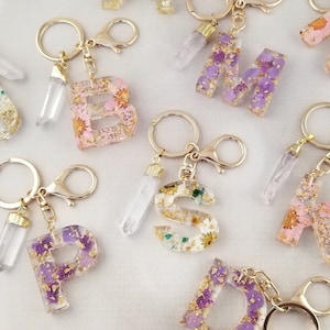 May include: A set of resin letter charms featuring dried flowers and gold flakes. Each charm is attached to a gold keyring with a clear quartz crystal pendant. The letters are B, M, S, K, D, and P.