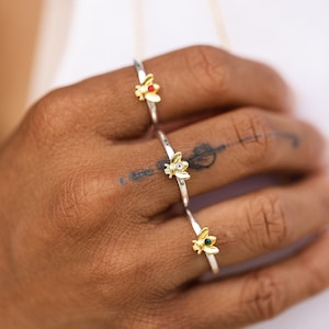 Pode incluir: Tr&ecirc;s an&eacute;is de tom prateado, cada um com um pequeno ornamento de abelha em tom dourado. A abelha superior tem uma pedra vermelha, a do meio uma pedra transparente e a inferior uma pedra verde. Os an&eacute;is s&atilde;o bandas finas e polidas.
