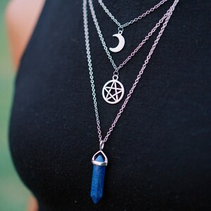 Pu&ograve; includere: Set di collane d'argento a strati con tre ciondoli: una luna crescente, un pentagramma e un cristallo blu. Le collane sono indossate sopra un top nero, creando un contrasto sorprendente. I ciondoli sono dettagliati e accattivanti.