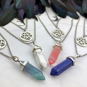 Pu&ograve; includere: Una collezione di collane argentate con pendenti di cristallo in vari colori, tra cui blu, bianco e rosa. Ogni collana presenta un ciondolo a pentagramma e a mezzaluna. Le collane sono esposte su una superficie bianca con piume scure sullo sfondo.