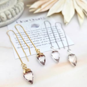 Puede incluir: Pendientes de cuarzo transparente con cadenas doradas y plateadas. Los pendientes presentan cristales de cuarzo transparentes y puntiagudos con una tapa dorada o plateada. El texto de la tarjeta dice "Clear Quartz is a powerful source of clarity."