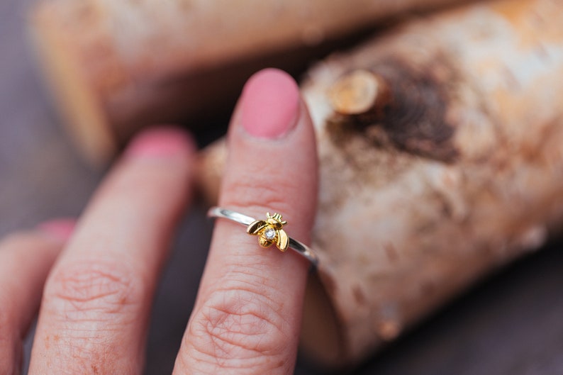 Pode incluir: Um anel de prata com um pequeno ornamento de abelha em tom dourado centrado na banda. A abelha tem uma min&uacute;scula pedra preciosa transparente de corte redondo incrustada no corpo, criando um ponto focal brilhante. A banda &eacute; fina e polida.
