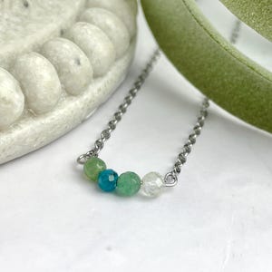 Może przedstawiać: Srebrny łańcuszek z pięcioma koralikami z kamieni szlachetnych. Koraliki są zielone, niebieskie, zielone, przezroczyste i zielone.