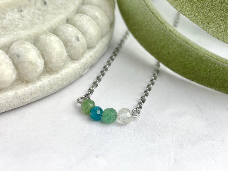 Może przedstawiać: Srebrny łańcuszek z pięcioma koralikami z kamieni szlachetnych. Koraliki są zielone, niebieskie, zielone, przezroczyste i zielone.