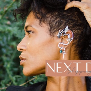May include: A silver dragon ear cuff with a green gemstone. The ear cuff is worn on the left ear of a person with curly hair. The background is a blurred image of green foliage.