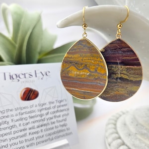 May include: Gold teardrop earrings with tiger's eye stone. The stones are a mix of brown, gold, and red with a swirling pattern. "Tigers Eye Power" is written on a card in the background.