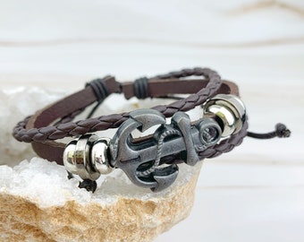 May include: A multi-strand bracelet featuring a dark brown leather cord and a braided dark brown leather strap. The centerpiece is a metallic silver-toned anchor charm with a rope detail, flanked by two polished silver-toned metal beads.