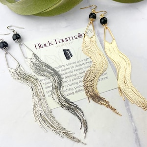 May include: Three pairs of long, dangling earrings with black gemstone beads and a fringe of gold, silver, and black chains. The earrings are displayed on a white surface with a card that reads "Black Tourmaline Protection".