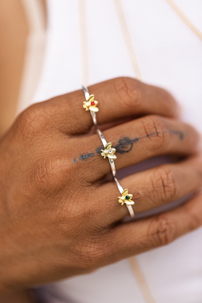 Pode incluir: Tr&ecirc;s an&eacute;is de tom prateado, cada um com um pequeno ornamento de abelha em tom dourado. A abelha superior tem uma pedra vermelha, a do meio uma pedra transparente e a inferior uma pedra verde. Os an&eacute;is s&atilde;o bandas finas e polidas.