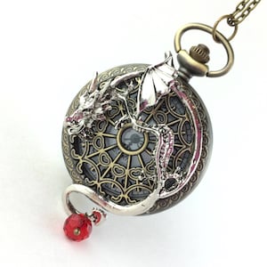 Puede incluir: Un colgante de reloj de bolsillo de dragón plateado con un dije de cristal rojo. La esfera del reloj tiene un diseño detallado y ornamentado.