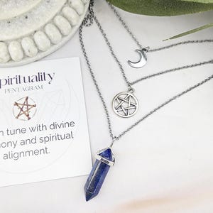 Pu&ograve; includere: Una collana d'argento a pi&ugrave; strati con tre ciondoli: una luna crescente, un pentagramma e una punta di cristallo blu. &Egrave; visibile anche una carta con il testo "Spirituality Pentagram" e la frase "Sono in sintonia con l'armonia divina e l'allineamento spirituale".