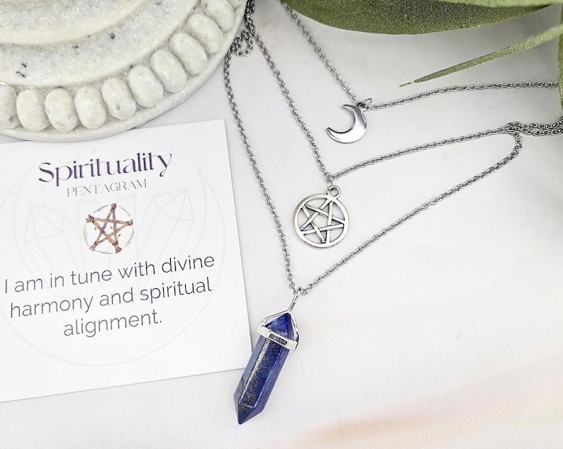 Pu&ograve; includere: Una collana d'argento a pi&ugrave; strati con tre ciondoli: una luna crescente, un pentagramma e una punta di cristallo blu. &Egrave; visibile anche una carta con il testo "Spirituality Pentagram" e la frase "Sono in sintonia con l'armonia divina e l'allineamento spirituale".