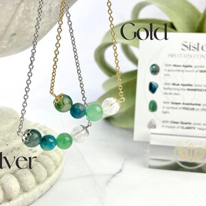 Może przedstawiać: Dwa naszyjniki z zielonymi, niebieskimi i przezroczystymi koralikami z kamieni szlachetnych. Naszyjniki leżą na białym tle z kartą, na kt&oacute;rej napisano "Sister Sisters Connection" i znajdują się słowa "Gold" i "Silver".