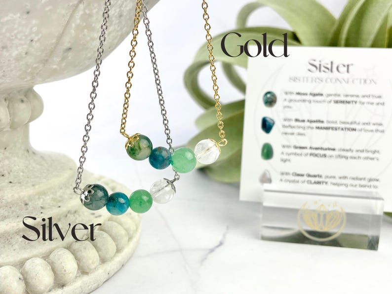 Może przedstawiać: Dwa naszyjniki z zielonymi, niebieskimi i przezroczystymi koralikami z kamieni szlachetnych. Naszyjniki leżą na białym tle z kartą, na kt&oacute;rej napisano "Sister Sisters Connection" i znajdują się słowa "Gold" i "Silver".