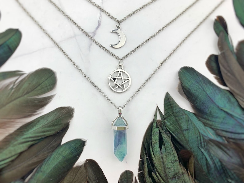Pu&ograve; includere: Un set di collane a strati con tre ciondoli: una luna crescente, una stella a cinque punte e un cristallo blu e verde. Le collane sono color argento e sono esposte su una superficie bianca con piume verdi.