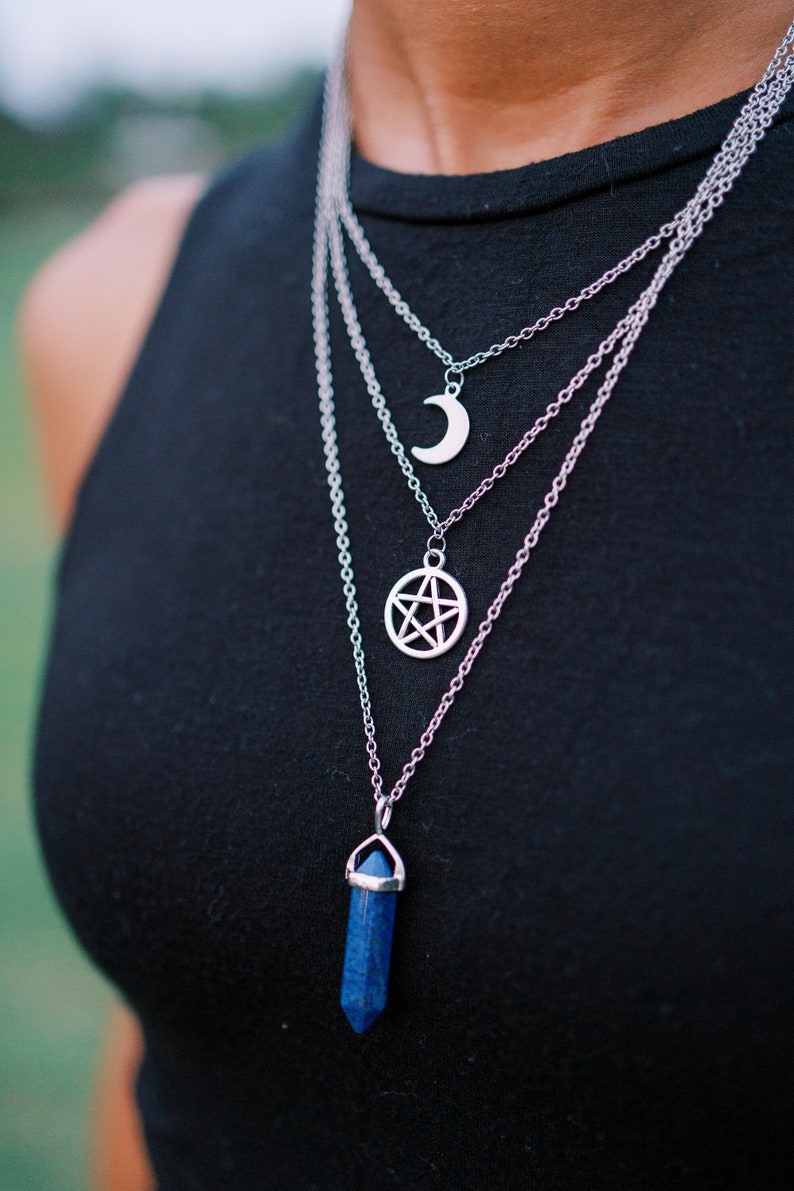Pu&ograve; includere: Set di collane d'argento a strati con tre ciondoli: una luna crescente, un pentagramma e un cristallo blu. Le collane sono indossate sopra un top nero, creando un contrasto sorprendente. I ciondoli sono dettagliati e accattivanti.