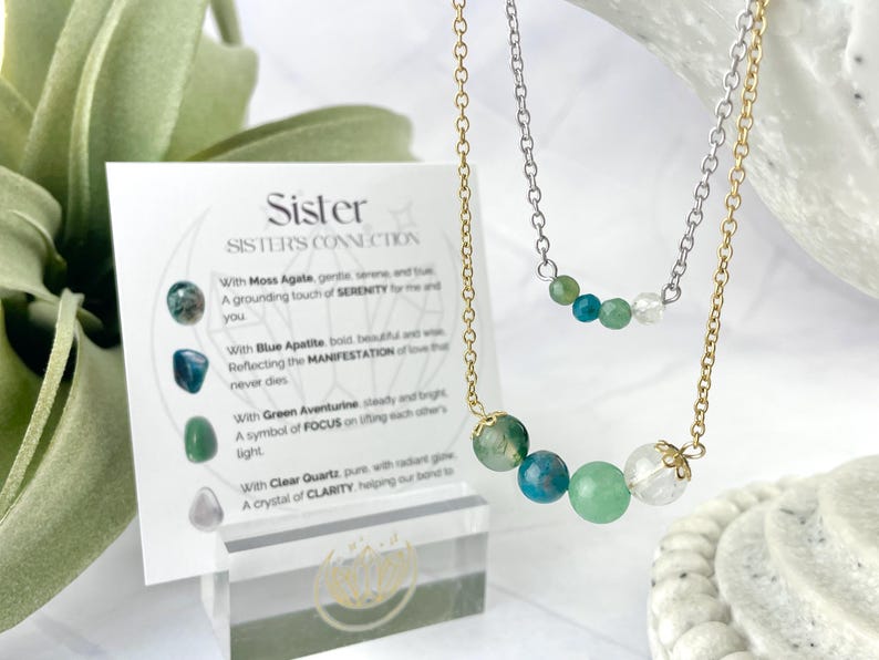 Może przedstawiać: Dwa złote naszyjniki z zielonymi i niebieskimi koralikami z kamieni szlachetnych. Naszyjniki są warstwowe i mają kartę z tekstem "Sister Sister's Connection" i opisami kamieni.