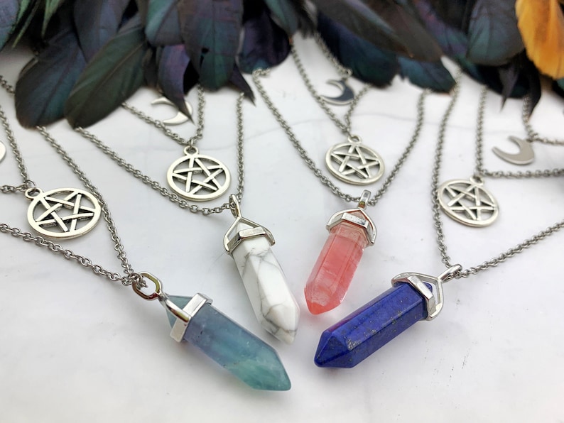Pu&ograve; includere: Una collezione di collane argentate con pendenti di cristallo in vari colori, tra cui blu, bianco e rosa. Ogni collana presenta un ciondolo a pentagramma e a mezzaluna. Le collane sono esposte su una superficie bianca con piume scure sullo sfondo.